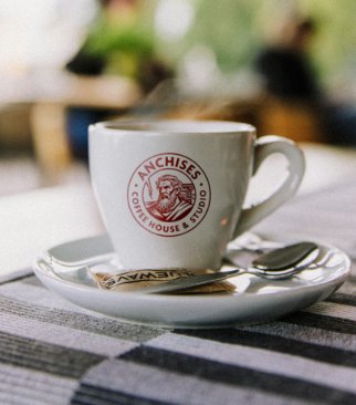 An espresso cup branded with the Anchises logo, resting on a saucer with a small spoon, on a striped grey tablecloth
